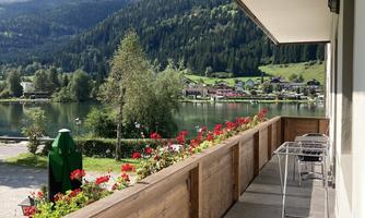 A balcony with a view of a beautiful landscape. In the foreground are chairs and tables as well as colorful flowers.