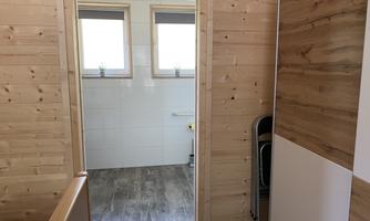 A bright room with wooden paneling and two windows. On the left side, a bathroom is visible.