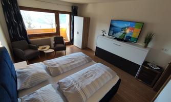 Bedroom with two beds, a television, and a comfortable seating area. Large windows provide a view of the outdoor terrace.