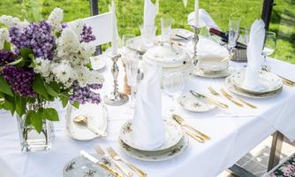 A beautifully set table outdoors with elegant plates and cutlery. In the center is a bouquet of lilacs and other flowers.