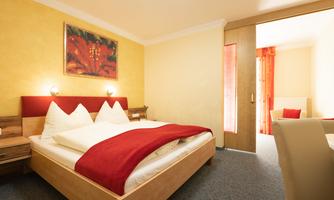 A bright bedroom with a double bed and red accents. A colorful painting hangs on one wall, and another room is visible through a door.