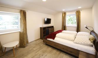A modern bedroom with a double bed, a TV, and a small table. The room has large windows with curtains and a bright, inviting style.