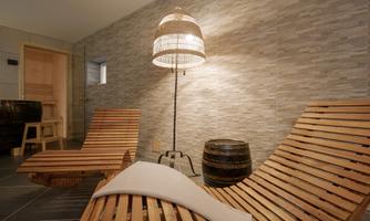 A cozy wellness area with wooden loungers and warm lighting. The walls are clad with modern tiles, and there is a sauna area in the background.