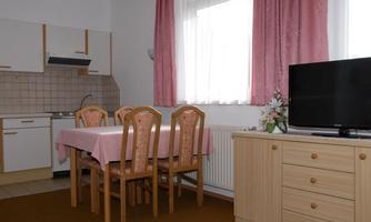 A bright kitchen with a dining area and a television. The table is covered with a pink tablecloth, and there are wooden chairs.