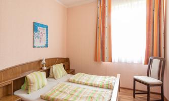 A simple room with two beds and light bedspreads. The room has orange curtains and a window that lets in natural light.