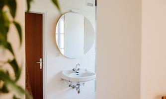 A modern bathroom with a round mirror and a sink. The walls are tiled in white, and a door leads to another room.