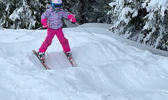 A girl in pink ski gear is skiing through the snow. Snowy trees can be seen in the background.