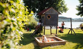 A playground with a wooden hut, swings, and a sandbox. In the background, there is a beautiful lake and green trees.