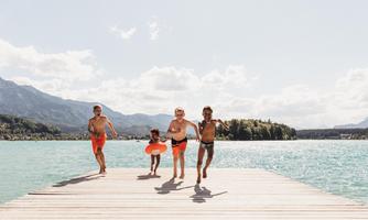 Four children are happily running on a dock by the water. In the background, mountains and a clear sky can be seen.
