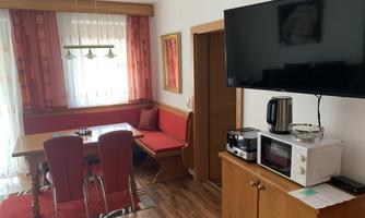 A cozy dining area with red chairs and a wooden bench. A television and kitchen appliances are placed on a sideboard.