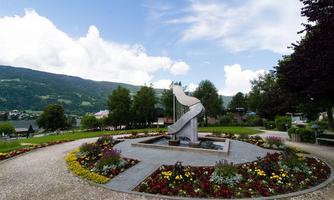 A beautiful park with a modern water feature and colorful flower beds. In the background, green hills and a clear sky can be seen.