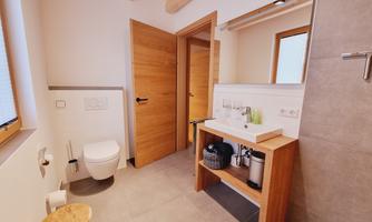 A modern bathroom with wood details and a large mirror. It includes a toilet, a sink, and a small stool.