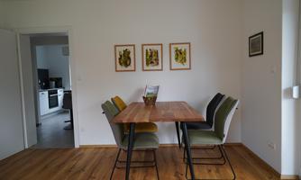 A dining area with a wooden table and colorful chairs. Three pictures are hung on the wall, and a modern kitchen is visible in the background.