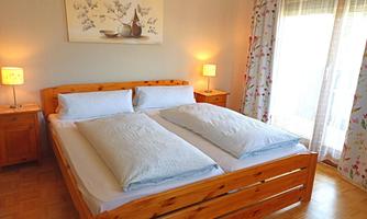 A cozy bedroom with a wooden bed and two mattresses. The room has soft light and a large French door.