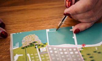 A hand holds a pen and points at a colorful map. The map depicts a stylized landscape with a lake and buildings.