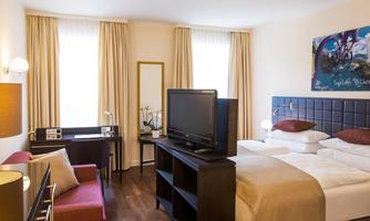 A modern hotel room with a double bed, a television, and a comfortable armchair. Large windows let in plenty of daylight.