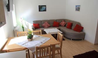 A cozy living room with a gray couch and red cushions. A wooden table with chairs and a flower decoration is also present.