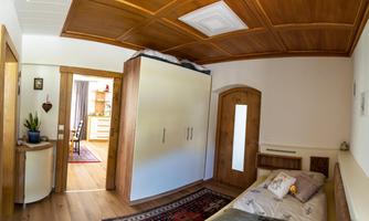 A cozy bedroom with a wooden ceiling panel and a large wardrobe. The room is bright and well-furnished, with a small table and a plant arrangement.