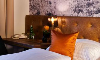 A cozy bedroom with an elegant headboard and an orange pillow. Next to the bed, there is a table with a bottle of water and a small flower.