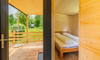 A bright wooden accommodation with one bedroom and a terrace. Outside, a green garden is visible.