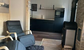 A cozy living space with two black reclining chairs and a wood stove. In the background, there is a modern kitchen with black cabinets.