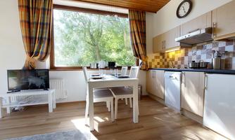 A bright kitchen with a large window and wooden ceiling. A dining table with white chairs and a television are also visible.