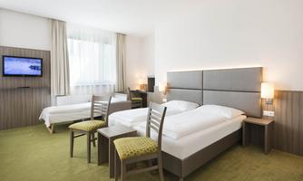 A modern hotel room with two beds, a television, and two chairs. The floor is covered with green carpet and there are large windows that let in a lot of light.