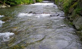A clear river flows between green banks. In the background, a small bridge can be seen.