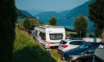 A parking space for motorhomes with a view of a lake and surrounding mountains. The landscape is green and sunny.