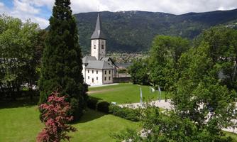 A picturesque landscape with a church and green trees. In the background, mountains and a blue sky are visible.