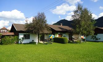 A cozy house with a green garden and beautiful trees. In the background, gentle hills and a blue sky can be seen.