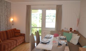A cozy living room with a striped couch and a dining area. Large windows offer a view of greenery.