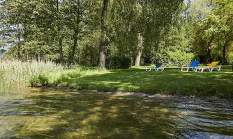 A quiet lake with a green shore and shade-providing trees. Two lounge chairs in blue and yellow invite you to relax.