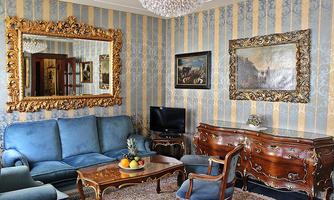 An elegant living room with blue sofas and a magnificent golden mirror. The walls are striped, and the furniture is antique and artistically decorated.