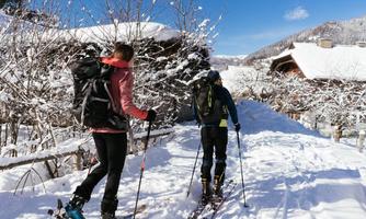 Two people are snowshoeing through a snowy landscape. In the background, there are snow-covered hills and some traditional huts.