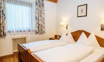 A cozy bedroom with a double bed and light furnishings. The windows let in plenty of natural light, and the decorative curtains create a pleasant atmosphere.