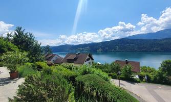 A picturesque landscape overlooking a clear lake and green hills. In the background, huts and a blue sky with white clouds can be seen.