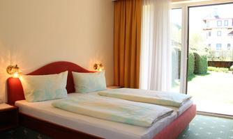 A cozy room with a double bed and two bright pillows. Large windows offer a view of a well-kept garden.