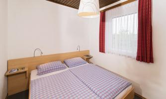 A cozy bedroom with a double bed and checkered bedspreads. Windows with curtains and a wooden ceiling create a homely atmosphere.