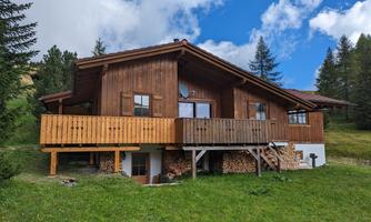 A cozy wooden house surrounded by green meadows and trees. The sky is blue with few clouds.
