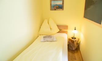 A simple bedroom with a single bed and yellow walls. A bedside lamp and a picture on the wall give the room a cozy atmosphere.