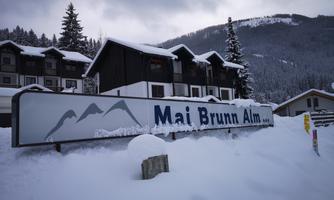 A snowy mountain area with the sign "Mai Brunn Alm". In the background, snow-covered trees and mountains are visible.