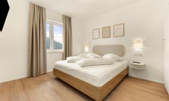 A bright bedroom with a double bed and modern furniture. The windows let in a lot of daylight, and there are decorative pictures on the wall.