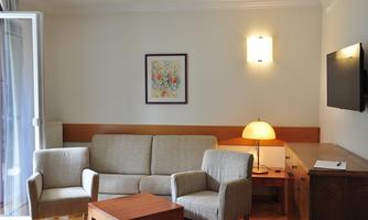 A bright living room with a comfortable sofa and two chairs. Next to a small table is a desk with a TV on the wall.
