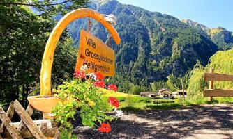 A sign reading "Villa Großglockner Heiligenblut" stands in front of a beautiful mountain landscape. In the foreground, colorful flowers can be seen, while the mountains rise majestically in the background.