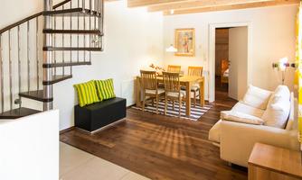 A modern living room with a bright wooden floor. There is a cozy seating area, a dining table, and a spiral staircase.