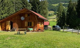A cozy wooden house surrounded by green meadows and tall trees. In the background, gentle hills and a blue sky can be seen.