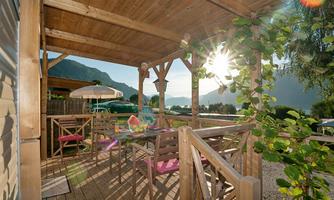 A cozy terrace with wooden chairs and a table, surrounded by plants. In the background, there are mountains and the sun is shining through the trees.