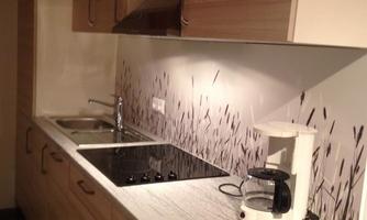 A modern kitchen with light wood cabinets and a gray countertop. It is equipped with a stove, a sink, and a coffee machine.