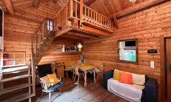 A cozy wooden house with a staircase and a rustic interior. The room is furnished with a dining table, chairs, and an elegant ceiling light.